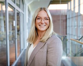Eine lächelnde Frau mit blonden Haaren und Brille trägt einen karierten Blazer. Sie steht in einem modernen Innenraum mit großen Fenstern.